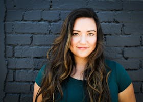 A captivating portrait of a woman smiling confidently in front of a brick wall, showcasing her long brunette hair and vibrant eyes.